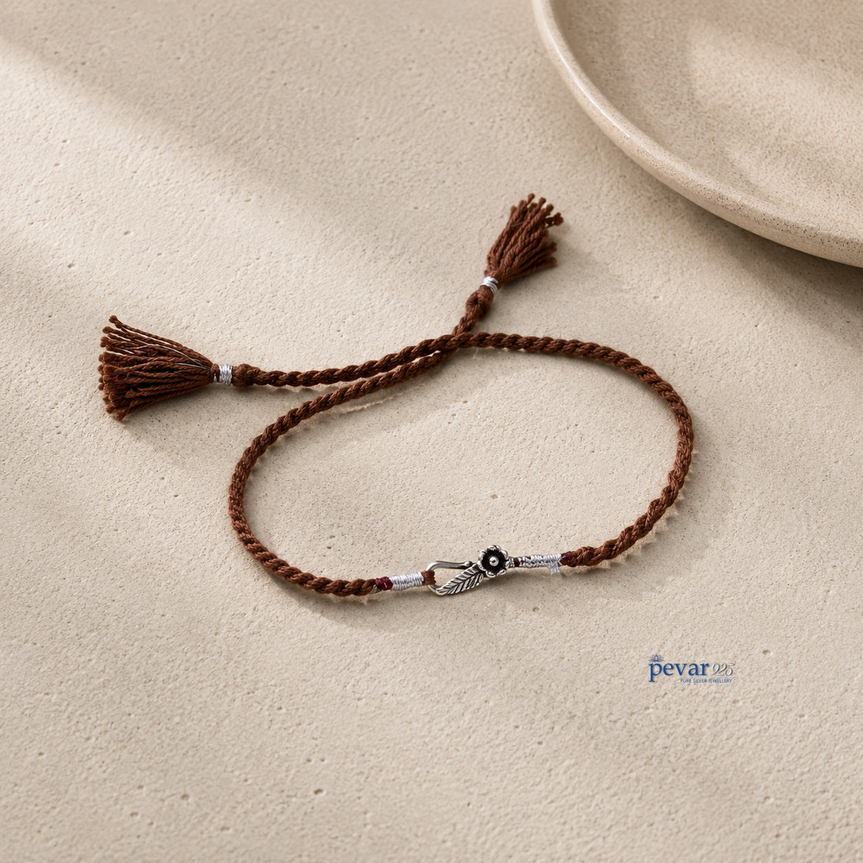 A brown braided cord bracelet with tassel ends and a silver-toned decorative clasp, displayed on a textured beige surface.