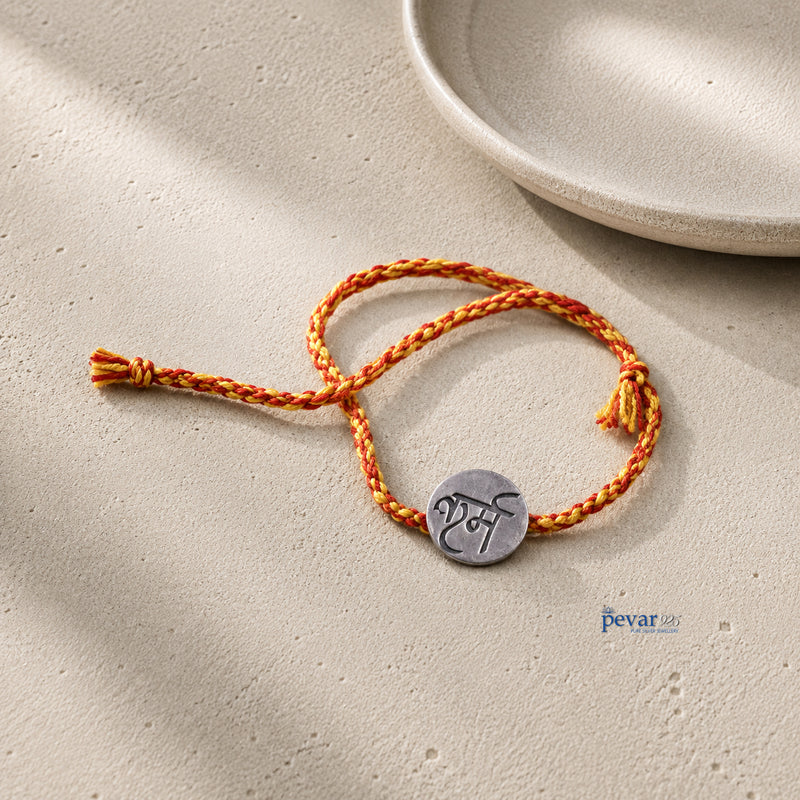 A braided orange and yellow cord bracelet with a round silver-toned metal charm engraved with a script, displayed on a textured beige surface with soft shadows.