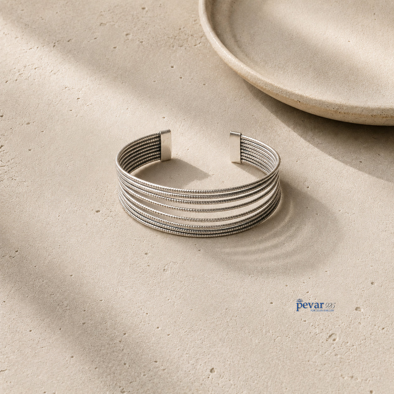 A silver-toned multi-strand cuff bracelet with a textured beaded pattern, displayed on a light beige stone surface with soft shadows and a ceramic plate in the background.