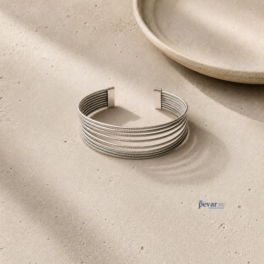 A silver-toned multi-strand cuff bracelet with a textured beaded pattern, displayed on a light beige stone surface with soft shadows and a ceramic plate in the background.