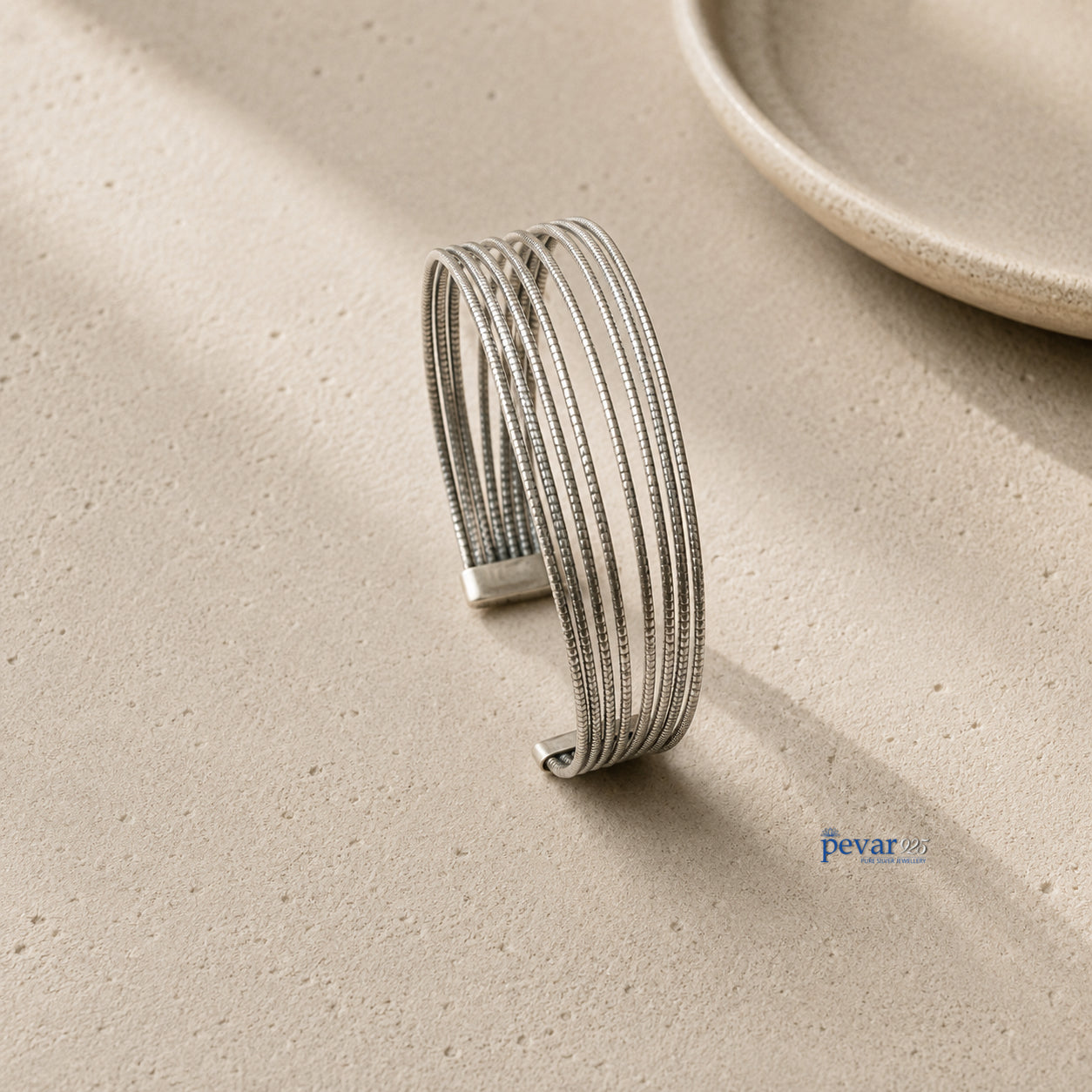 A silver-toned multi-strand cuff bracelet with textured details, displayed on a textured beige surface with soft shadows.