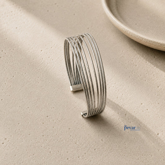 A silver-toned multi-strand cuff bracelet with textured details, displayed on a textured beige surface with soft shadows.