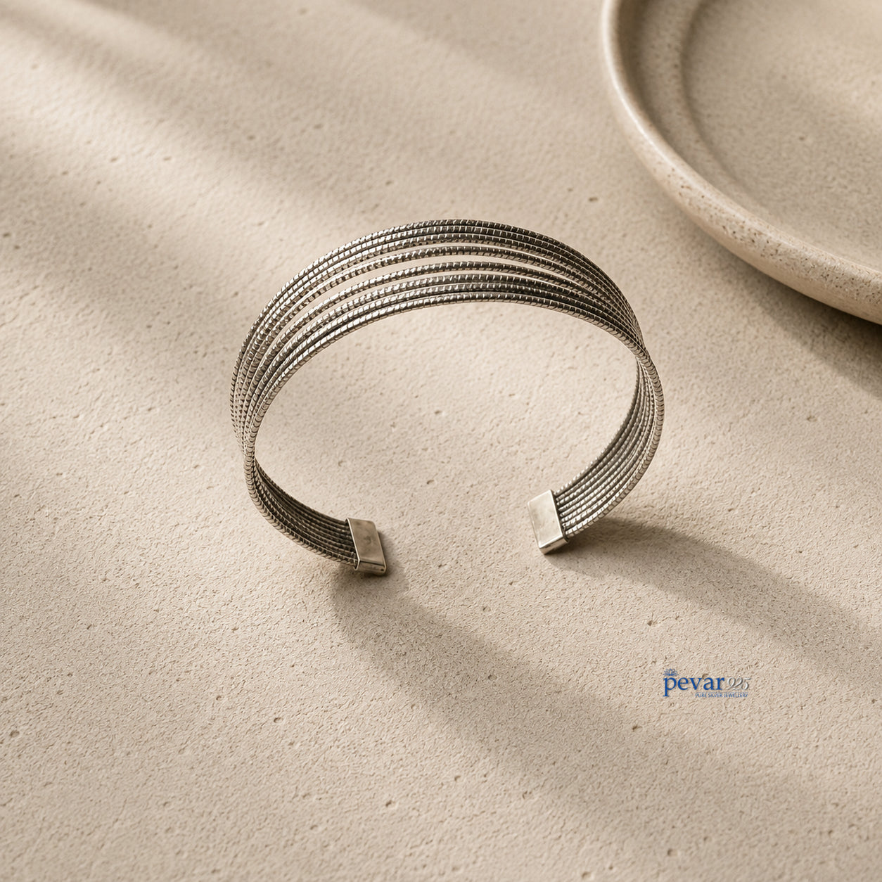 A silver-toned open cuff bracelet with a textured, multi-strand mesh-like design, displayed on a light beige textured surface with soft shadows.