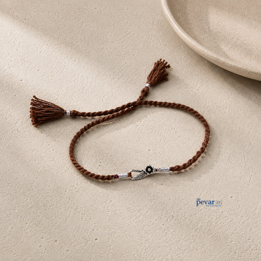 A brown braided cord bracelet with tassel ends and a silver-toned decorative clasp, displayed on a textured beige surface.
