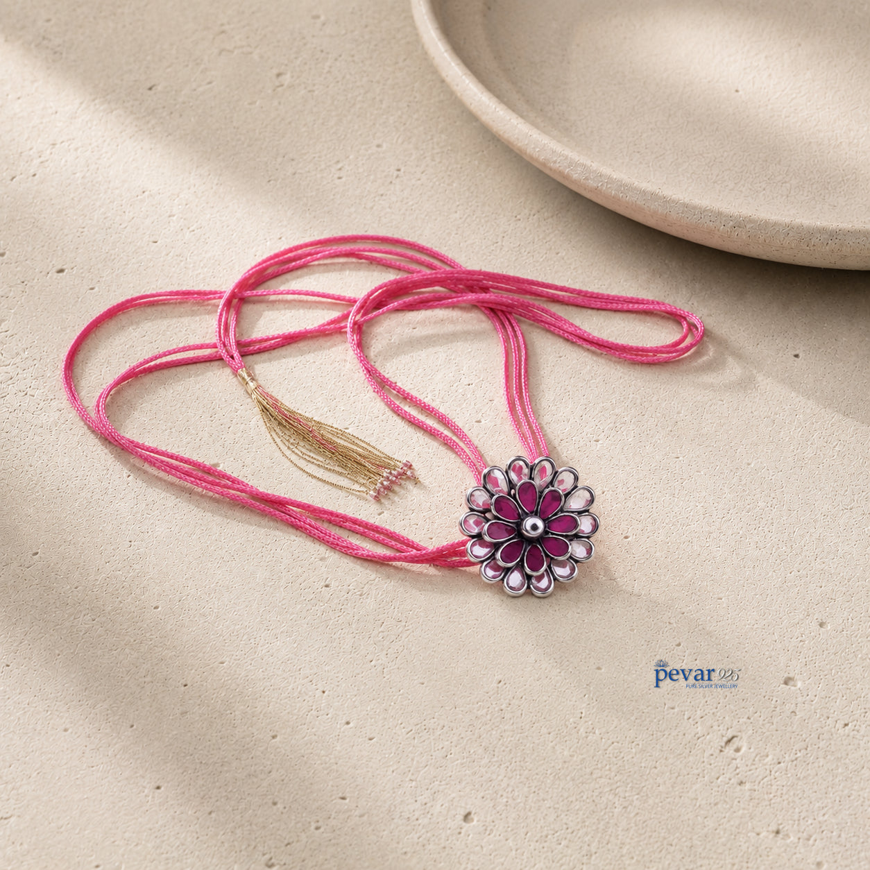 A pink thread necklace with a silver-toned flower pendant featuring pink and white enamel-like detailing, resting on a textured beige surface with a small tassel accent.