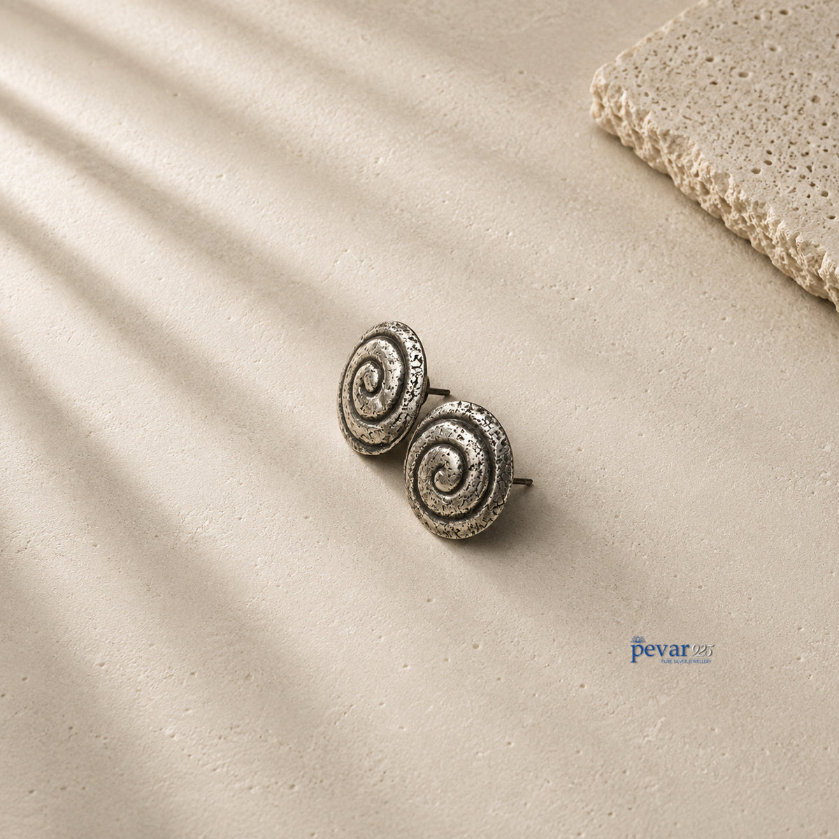 A pair of silver spiral stud earrings with a textured, oxidized finish, displayed on a beige textured surface with soft diagonal shadows and a stone prop in the corner.
