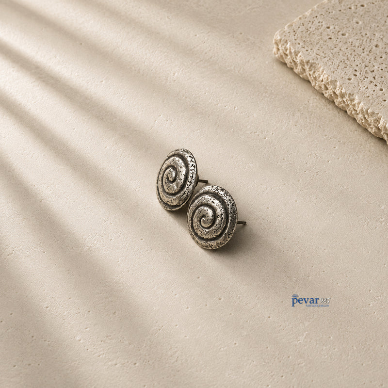 A pair of silver spiral stud earrings with a textured, oxidized finish, displayed on a beige textured surface with soft diagonal shadows and a stone prop in the corner.