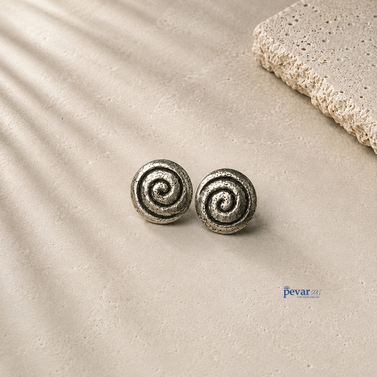 A pair of round spiral stud earrings in a textured silver-tone metal, displayed on a light beige stone surface with soft leaf shadows and a decorative stone slab in the corner.