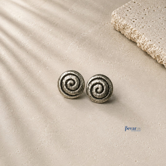 A pair of round spiral stud earrings in a textured silver-tone metal, displayed on a light beige stone surface with soft leaf shadows and a decorative stone slab in the corner.