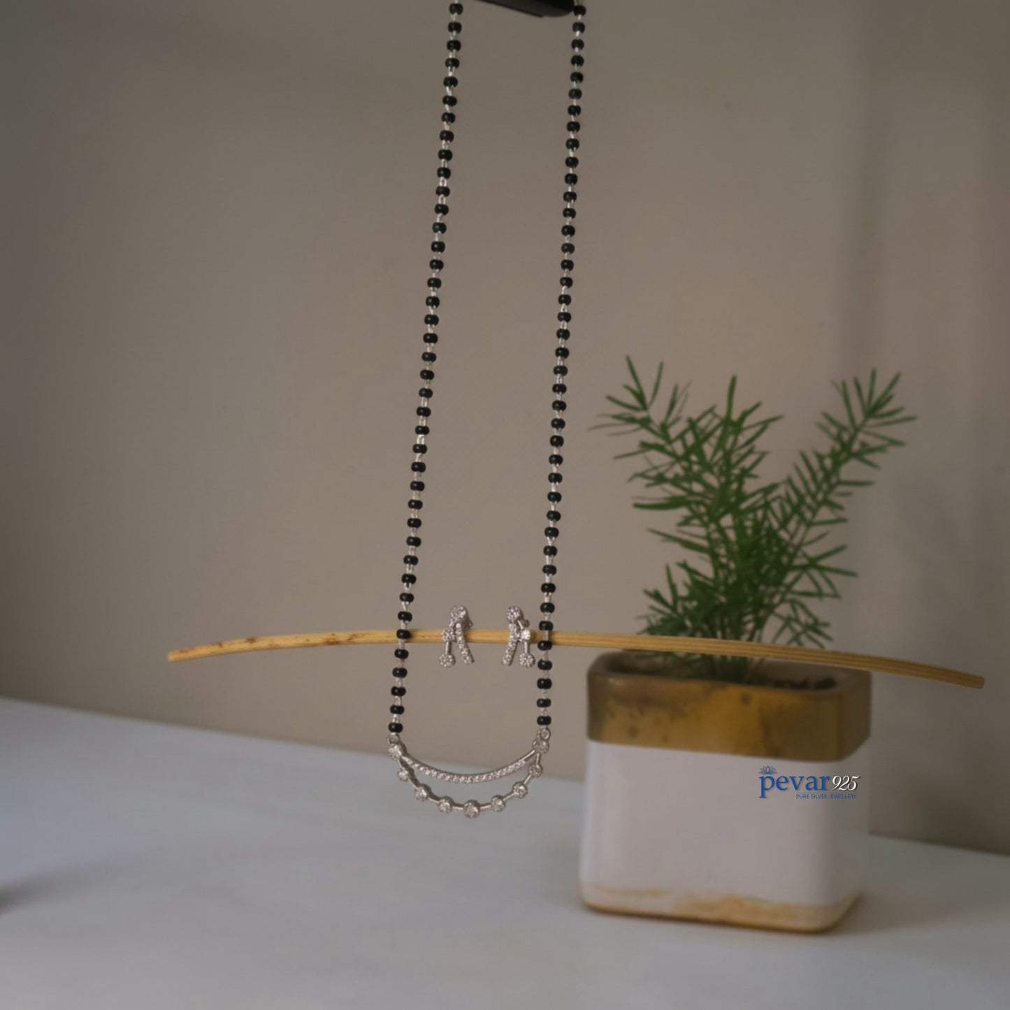 Necklace with chain detail on a stick next to a plant in a pot with 'pevar' branding.
