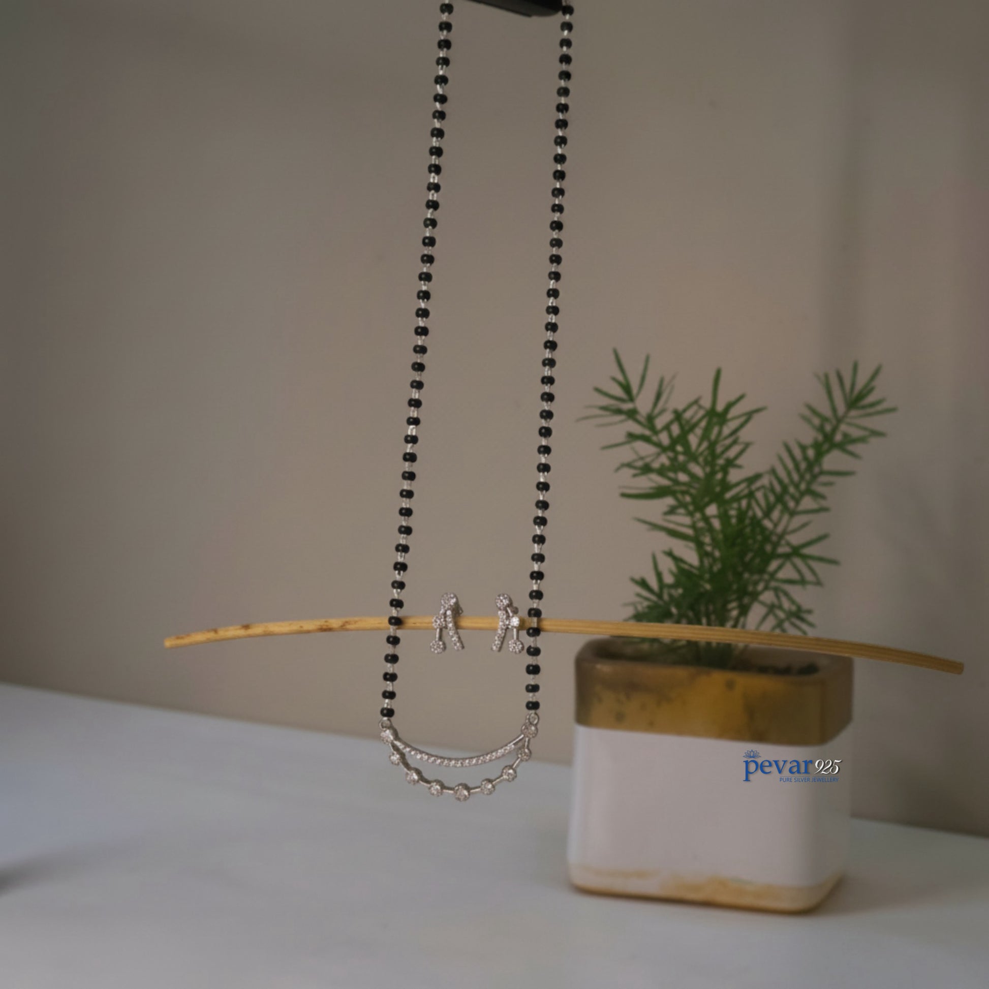 Necklace with chain detail on a stick next to a plant in a pot with 'pevar' branding.