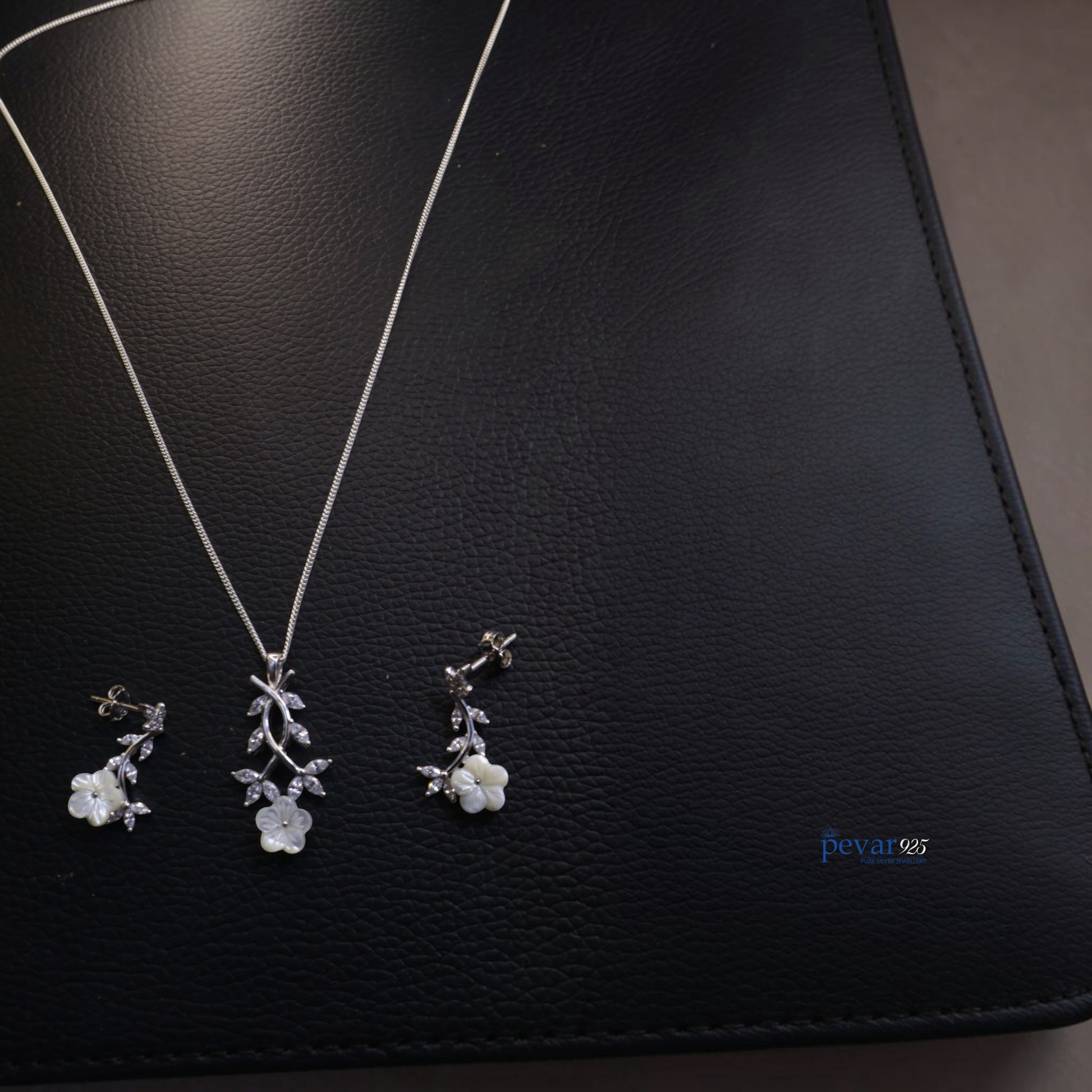 Silver floral necklace and earrings on a black leather surface with 'pevar' branding.