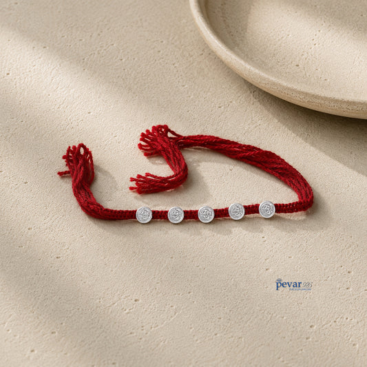 A red braided cord bracelet with five small silver circular charm beads, laid flat on a textured beige surface with soft shadows and a ceramic plate edge in the background.